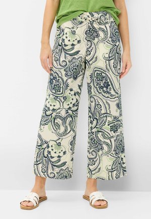 Person trägt beige weite Hose mit blau-grünem floralem Paisley-Muster, grünes Oberteil und weiße Slip-On-Sandalen.