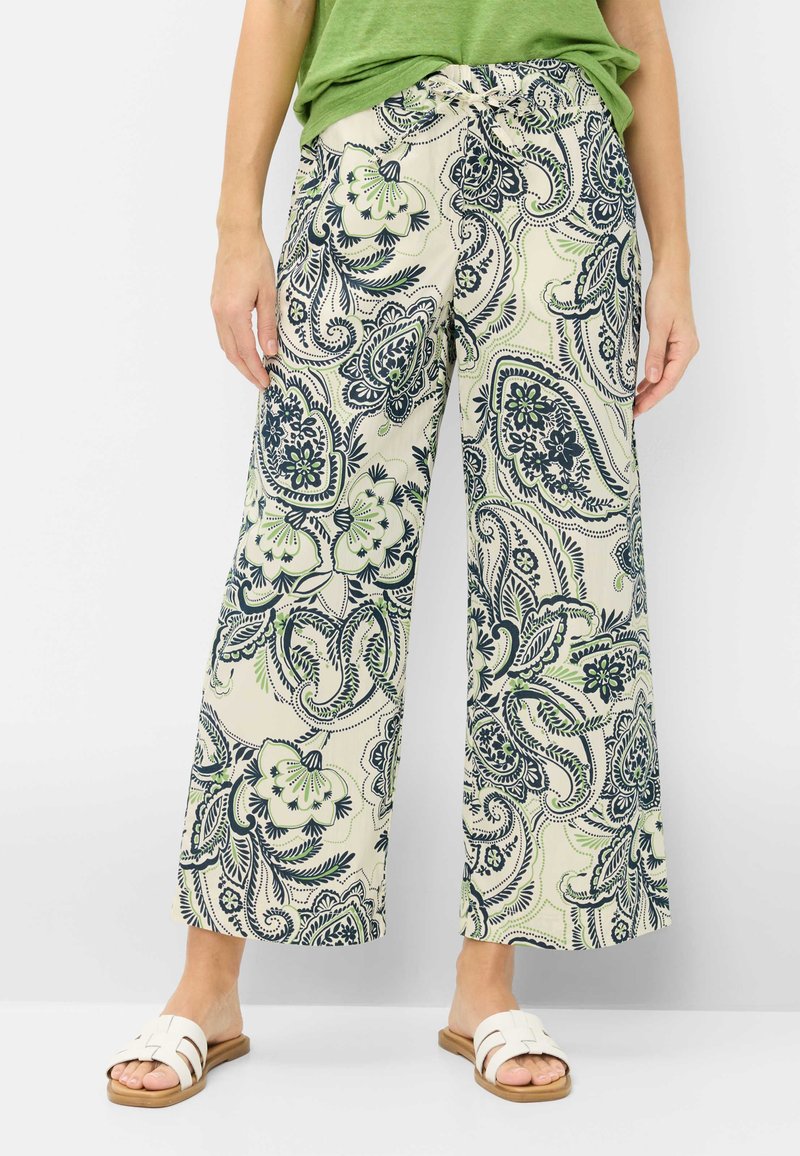 Personne portant un pantalon beige à jambes larges avec un imprimé paisley floral bleu et vert, un haut vert et des sandales blanches à enfiler.