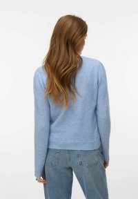 Kobieta o długich brązowych włosach, ubrana w jasnoniebieski sweter i niebieskie dżinsy, stojąca tyłem do kamery na białym tle.