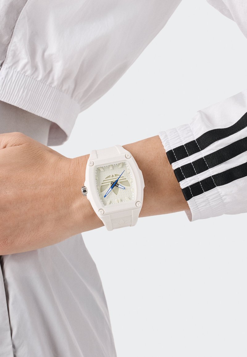 Orologio in gomma bianca con un quadrante quadrato, dial crema testurizzato, lancette blu e dettagli neri. Indossato su un polso con una giacca bianca con strisce nere.