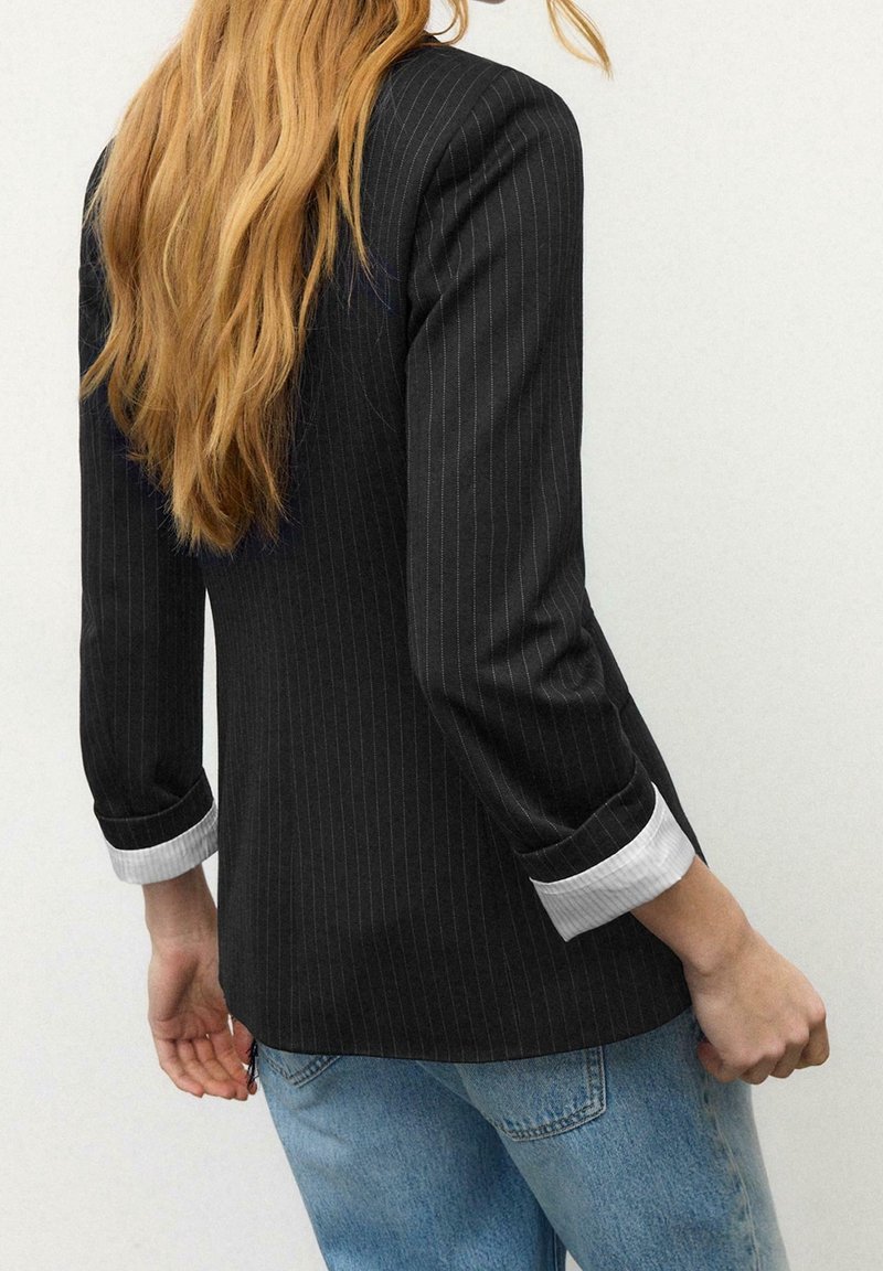 Femme aux cheveux longs portant un blazer noir à fines rayures avec des poignets blancs et un jean bleu, debout devant un fond clair uni.