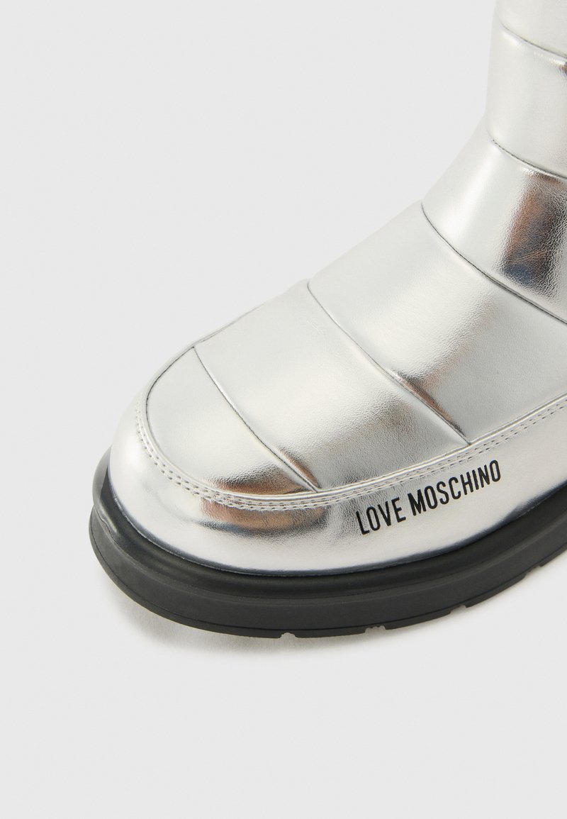 Bottine métallique argentée avec un design rembourré, bout arrondi, semelle en caoutchouc noire et marquage "LOVE MOSCHINO" sur le côté.