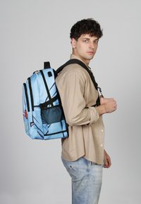 Mochila azul con detalles en negro, múltiples compartimentos y un diseño de dibujos animados. El modelo la lleva sobre un hombro frente a un fondo liso.