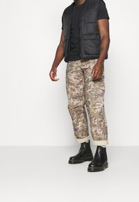 Svart vadderad väst över en svart t-shirt, ihopparad med kamouflagefärgade cargobyxor och svarta ankelboots med gula sömmar.
