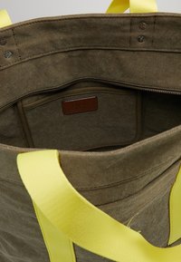 Canvas tote bag i khaki med två ljusa gula handtag, som har en dragkedjeficka och en läderetikett inuti.