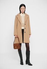 Abrigo de lana beige con cuello muesca, botones negros, combinado con un top de punto color crema, bolso marrón, pantalones oscuros y botas negras hasta la rodilla.