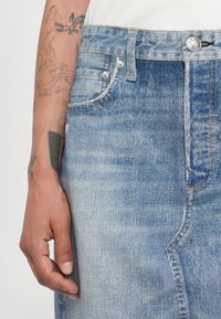 Gonna in denim azzurro chiaro con vita alta, tasca anteriore e cuciture a contrasto. Texture liscia con leggere imperfezioni che suggeriscono usura.