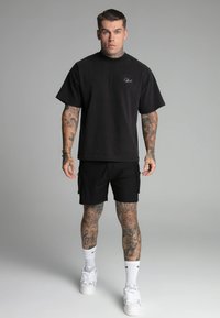 Camiseta de algodón negra con mangas cortas, exhibida con pantalones cortos cargo negros. Presenta tatuajes en los brazos y piernas, zapatillas blancas y calcetines de caña corta.