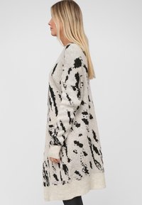 Sweter sukienka w kolorze beżowym z czarnymi abstrakcyjnymi wzorami, z długimi rękawami, luźnym krojem oraz ściągaczami przy rękawach i u dołu.