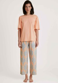 Perzik kleurige top met korte mouwen en geplooide schouders, gecombineerd met lichtoranje broek met een blauw bloemmotief en wijde pijpen.