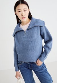 Lys blå ribbet genser med glidelås og vid krage, båret over en rosa topp, sammen med blå denimjeans.
