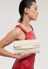 Beige Leder-Clutch mit Überklapp-Design, ausgestattet mit einem metallischen Verschluss, gehalten in einer Hand vor einem roten Lederoutfit. Glatte Textur.