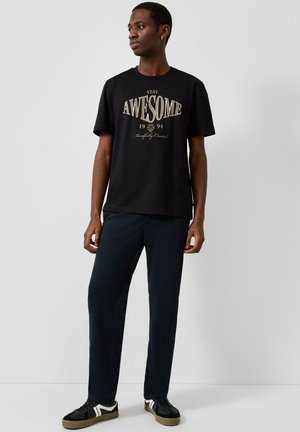 Giovane uomo che indossa una maglietta nera con il testo "Stay Awesome 1994", pantaloni scuri e scarpe da ginnastica nere con strisce bianche, in piedi davanti a una parete liscia.