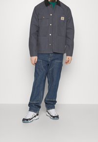Giacca da lavoro grigia con quattro tasche frontali, bottoni dorati, colletto nero, abbinata a jeans blu e sneakers bianche con dettagli neri.