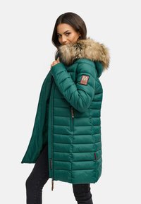 Chaqueta acolchada verde con capucha forrada de piel, textura acolchada, bolsillos laterales y un parche de logo en la manga. Cierre frontal con cremallera.