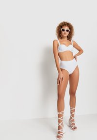 Balta bikini komplekts ar strukturētu krūštura augšdaļu un augsto jostasvietu apakšdaļām. Modeli papildina baltas siksniņu kurpes un saulesbrilles, ar neitrālu fona ainu.