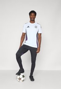 Hvit fotballdrakt med lilla og grå detaljer, svarte treningsbukser, og svarte fotballsko; modellen står på en fotball.