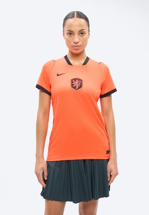 Femme portant un maillot de sport orange à manches courtes avec des bordures noires et un emblème de lion, associé à une jupe plissée foncée.