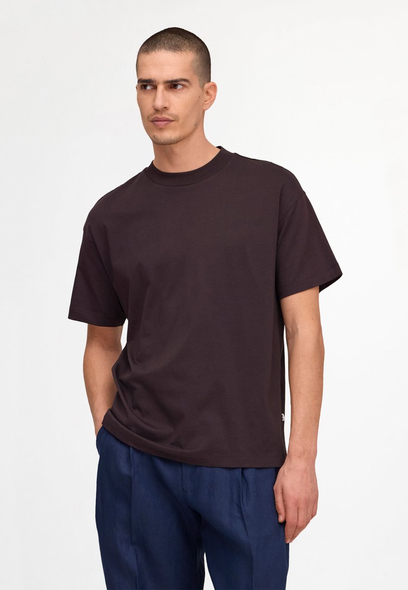 Homme aux cheveux courts portant un T-shirt marron foncé à manches courtes et un pantalon bleu, regardant sur le côté avec une main dans la poche.