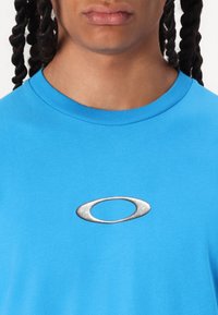 Svijetloplavi pamučni majica s srebrnim ovalnim logom u centru prsnog dijela. Okrugli izrez s rebrastim rubom, glatke teksture.