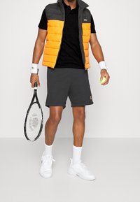 Pufferväst i orange och grå, svart polo, grå shorts, vita sportskor, håller en tennisracket och boll.