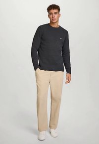 Mørkegrå strikkegenser med rund hals, ribbekanter ved ermer og nederkant, kombinert med lysebeige bukser og hvite joggesko, stående mot en nøytral bakgrunn.