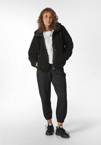 Giacca in pile nera con zip e cappuccio regolabile, indossata sopra una t-shirt bianca, abbinata a pantaloni jogger scuri e sneakers nere.