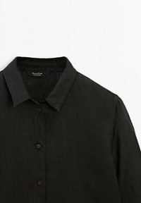 Chemise en lin noire avec col à boutons, présentant un tissu texturé et des boutons noirs le long de la patte de boutonnage avant.
