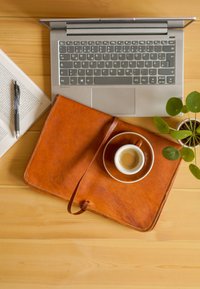 Bruin lederen notitieboek met een strik, een witte koffiekop op een schotel, een laptop, een pen, een boek en een kleine groene plant op een houten oppervlak.