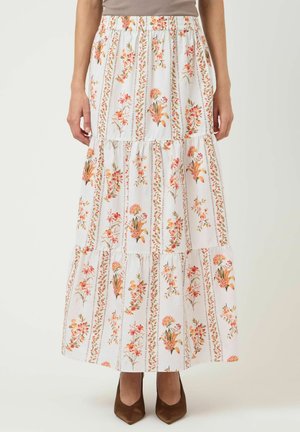 Kvinne som har på seg et langt hvitt skjørt med vertikale blomstrede striper i oransje og grønt, kombinert med brune pumps med spiss tå.