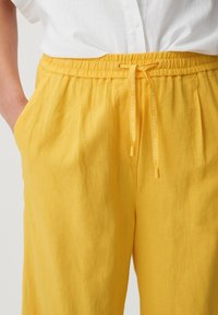 Geel linnen broek met een elastische tailleband en een trekkoord. Aan beide zijkanten zijn zakken zichtbaar, met een gestructureerd oppervlak.