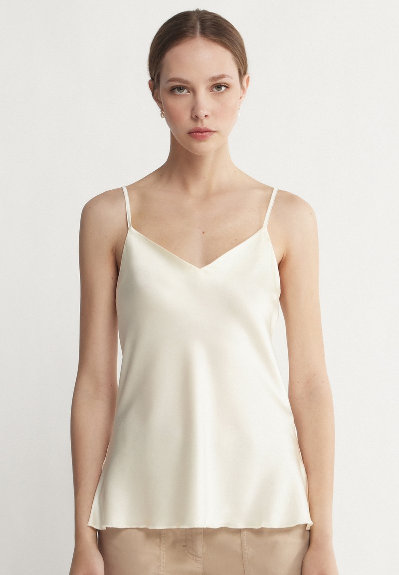 Krämfärgad satinkjol med justerbara tunna axelremmar, V-urringning och en svängande nederkant som mjukt hänger över midjan. Slät, glansig textur.