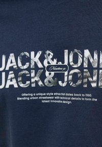Marineblå bomulds T-shirt med fed hvid typografi; teksten inkluderer "JACK & JONE" og "Nineteen 90" med en struktureret effekt.