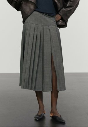 Femme portant une jupe plissée grise avec une fente à l'avant, un haut noir, une veste en cuir marron et des chaussures slip-on noires, debout sur un sol gris.