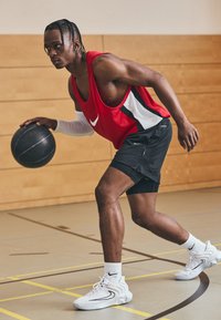 Camiseta sin mangas roja de Nike con detalles en negro y blanco, pantalones cortos negros y calzado blanco. Cancha de baloncesto interior con paredes de madera y marcas en el suelo.