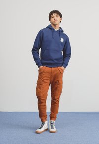 Temno modra kapuca z sprednjim žepom in logotipom, v kombinaciji z oranžnimi cargo hlačami, ki vsebujejo žepe. Obuta z belimi in rjavimi supergami na višji peti.