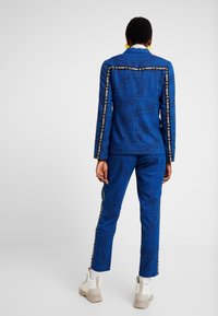 Costume à carreaux bleu en tissu avec des accents de texte noir. Comprend une veste cintrée et un pantalon ajusté, complétés par une bande métallique.