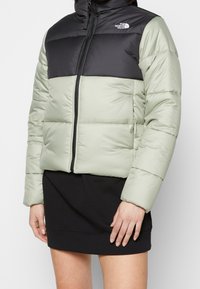 Padded jacket in light green and black, featuring a high collar, zip closure, and elasticized cuffs. The material has a smooth finish.