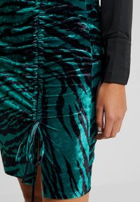 Grön och svart tigerstripad sammet klänning med rynkdetaill, med en dragsko i fållen. Närbild av tygtextur och mönster.