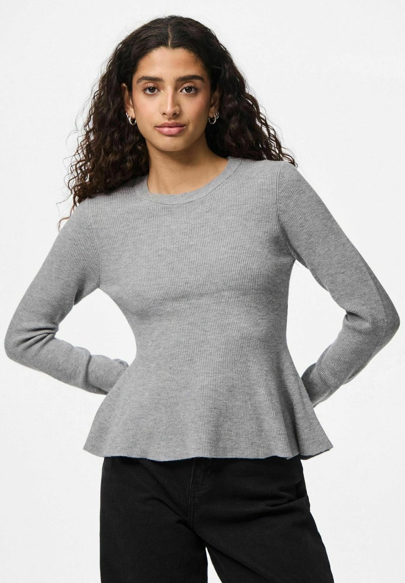 Jeune femme aux cheveux noirs bouclés portant des boucles d'oreilles en argent, un pull gris ajusté à manches longues et un pantalon noir, se tenant les mains sur les hanches.