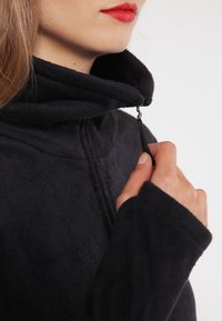 Mustapeltilakki, jossa on korkea kaulus. Takissa on vetoketjukiinnitys kaulalla ja pitkät hihat lisälämpöä ja mukavuutta varten.