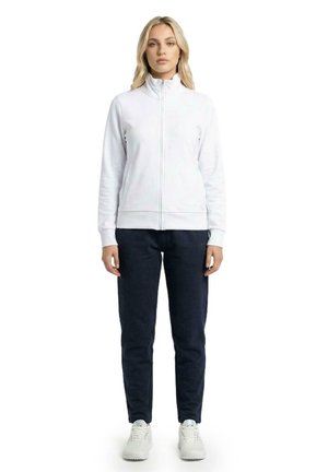 Une femme porte une veste blanche zippée, un pantalon foncé et des baskets blanches sur un fond uni.