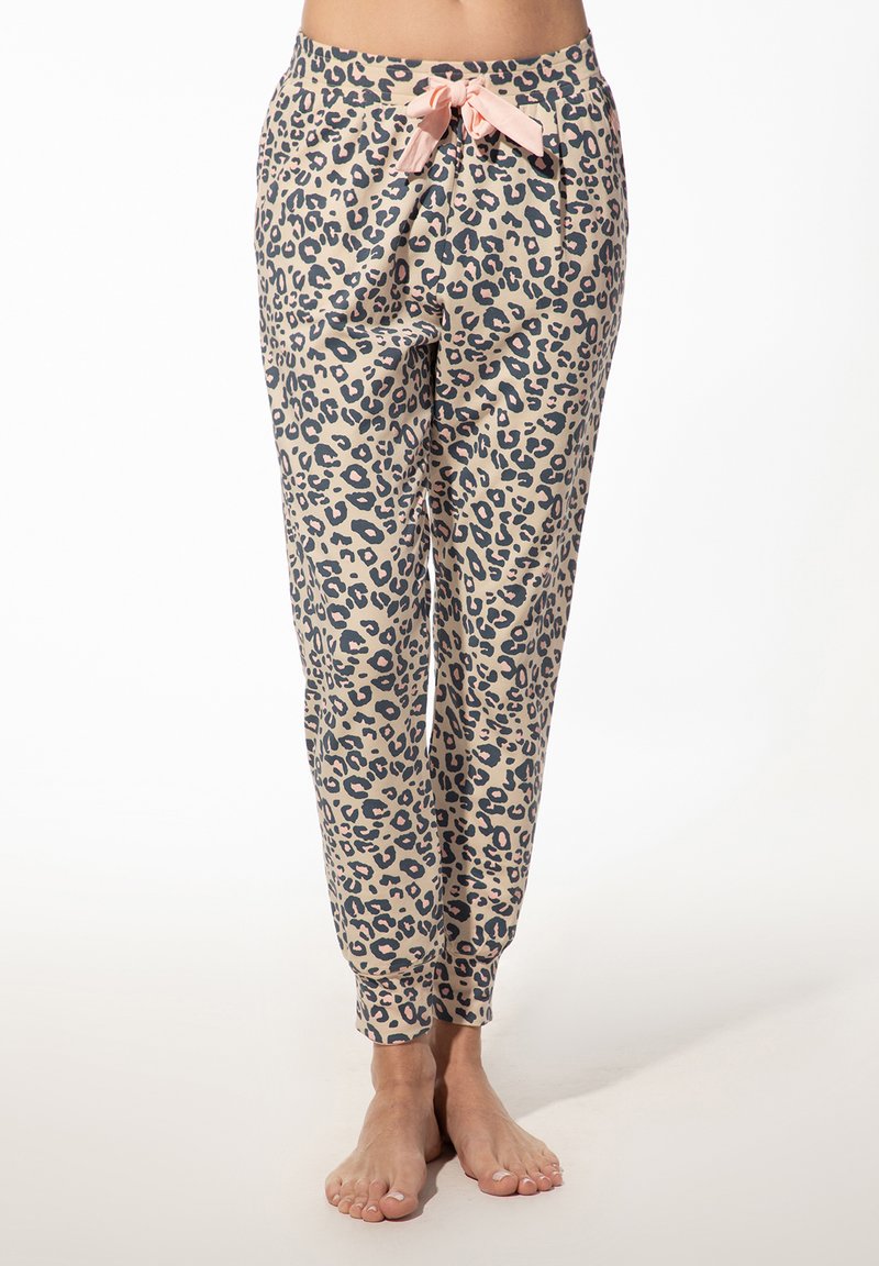 Vrouw in beige pyjamabroek met luipaardprint en een roze tailleband, blootvoets staand op een lichte vloer tegen een witte achtergrond.