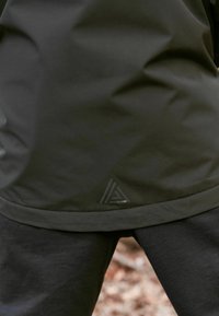 Nærbillede af en person iført en sort jakke og bukser, stående udendørs på en jord dækket af blade.