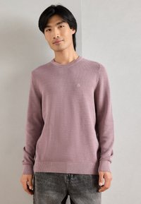 Lyse lilla sweater med tekstureret strik, rund halsudskæring og ribbet kant. Den har et lille logo på brystet og lange ærmer.