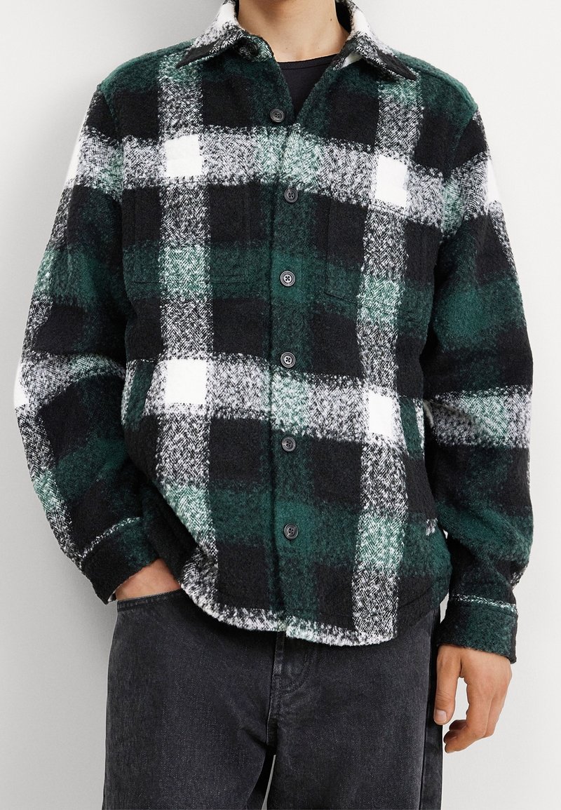 Mand iført en knappet skjortejakke i uld med mørkegrøn, sort og hvid tern, sammen med sorte bukser, stående med den ene hånd i lommen.