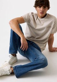 Beigā kokvilnas t-kreklā ar logo, zilās denim biksēs un baltos sporta apavos. Modelis sēž, demonstrējot brīvu piegriezumu un ikdienišķa stila elementus.