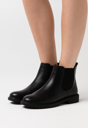 Bottes Chelsea en cuir noir montant jusqu'à la cheville, avec des panneaux latéraux élastiques et des semelles striées, portées sur jambes nues sur un fond neutre.