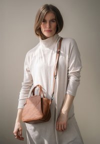 Braune Lederhandtasche mit zwei Griffen und einem Umhängeriemen, mit einer glatten Textur und einem schrägen Design. Getragen mit einem hellen cremefarbenen Rollkragenkleid.