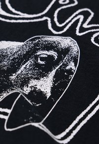 Μαύρο T-shirt με λευκή γραφική εκτύπωση προσώπου, με υφή λεπτομέρειας και καμπυλωτά γράμματα πάνω από το σχέδιο.